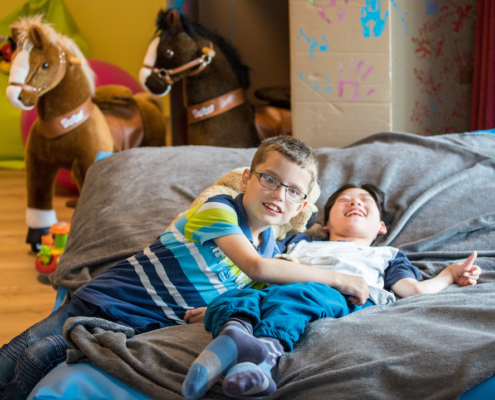 Zwei Jungen mit Beeinträchtigung liegen auf einem großen Kissen und grinsen in Richtung Kamera. Im Hintergrund ist Spielmaterial zu sehen.